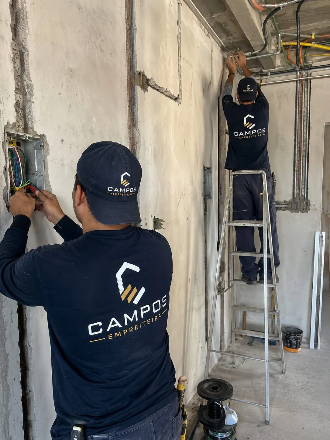 Equipe da Campos Empreiteira executando instalação elétrica em obra, com profissional em escada ao fundo